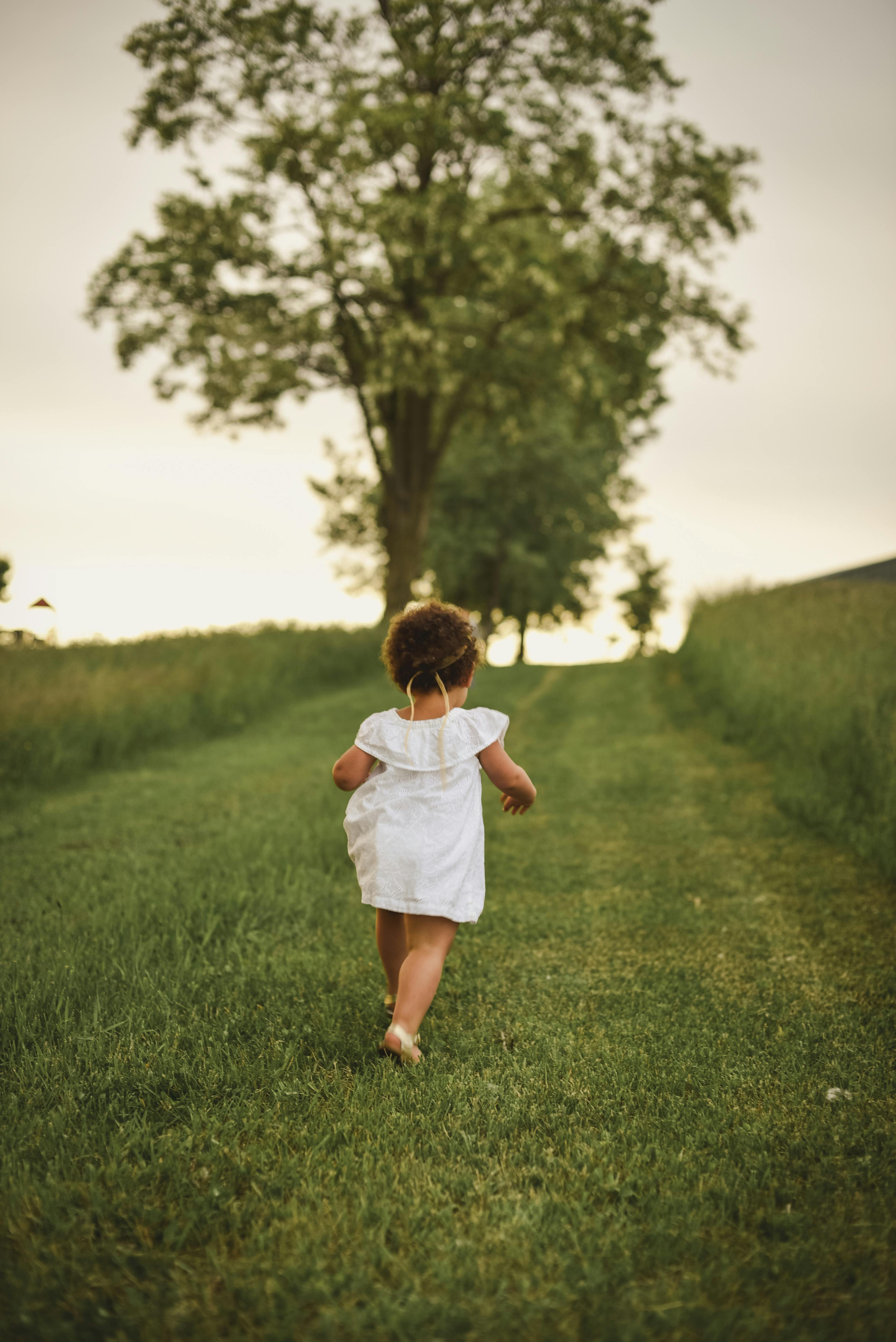 Child in a white dress running through a grassy field with a tree in the background