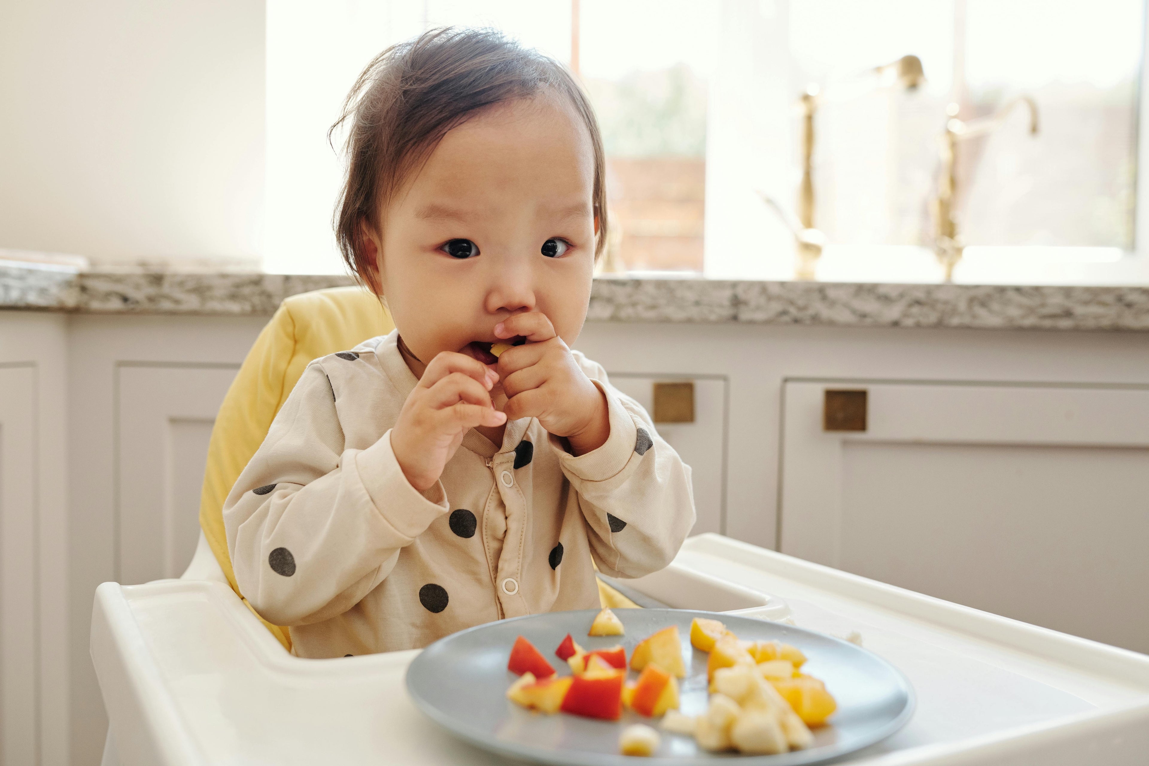 Child sitting in a high chair eating fruit at a kitchen table.